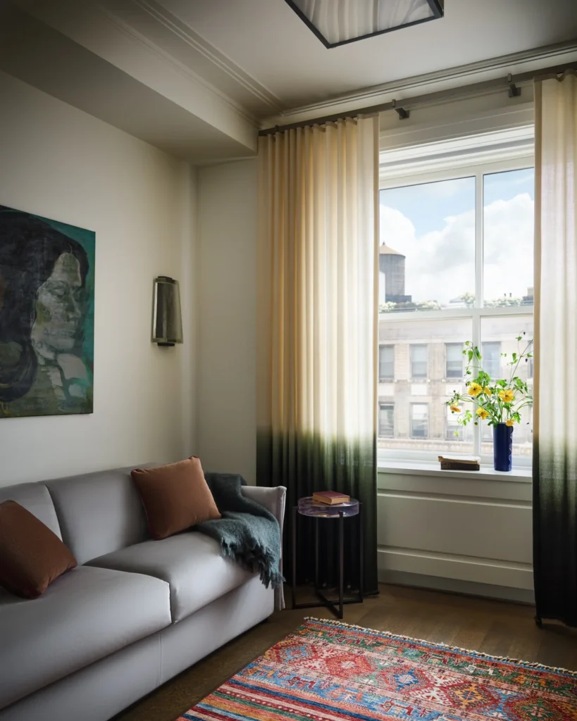 Sitting room with a gray sofa, rust pillows, colorful rug and cream curtains fading to dark green.