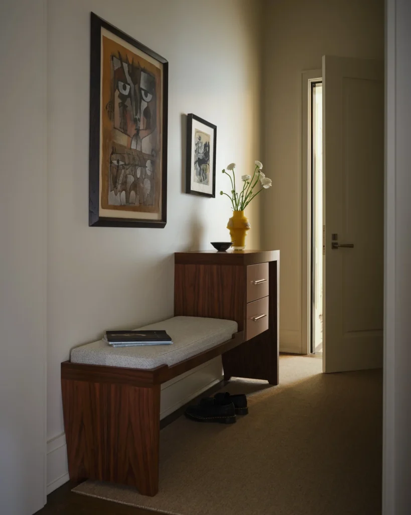 Belnord hallway with a golden stone pedestal, amber vase of greenery and wood bench beneath framed art.