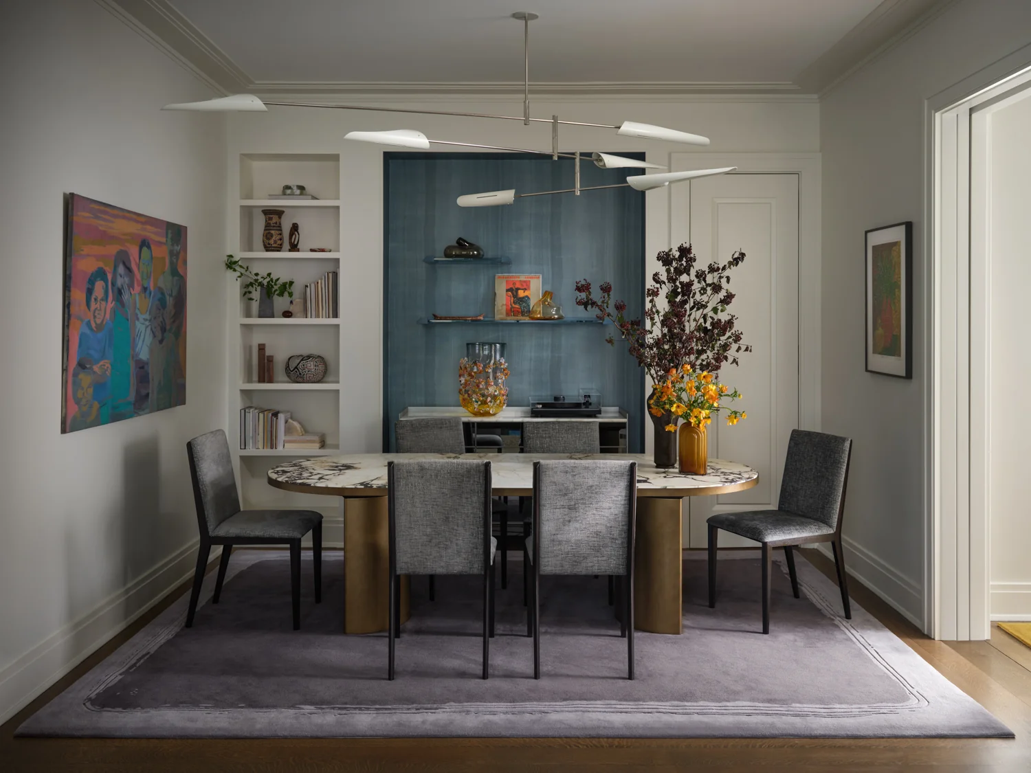 Dining room with a marble table, gray chairs, branching chandelier and blue display shelves filled with art and objects.
