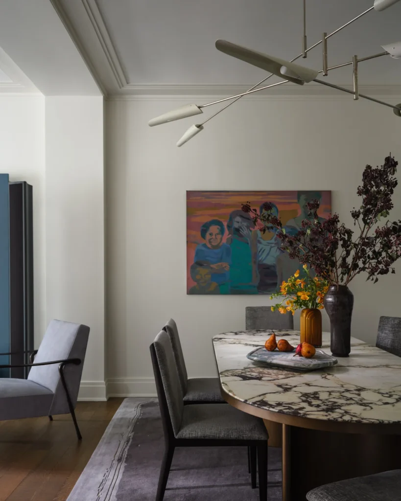 Veined marble dining table with sculptural vases, yellow flowers and a fruit tray beneath colorful portrait art.