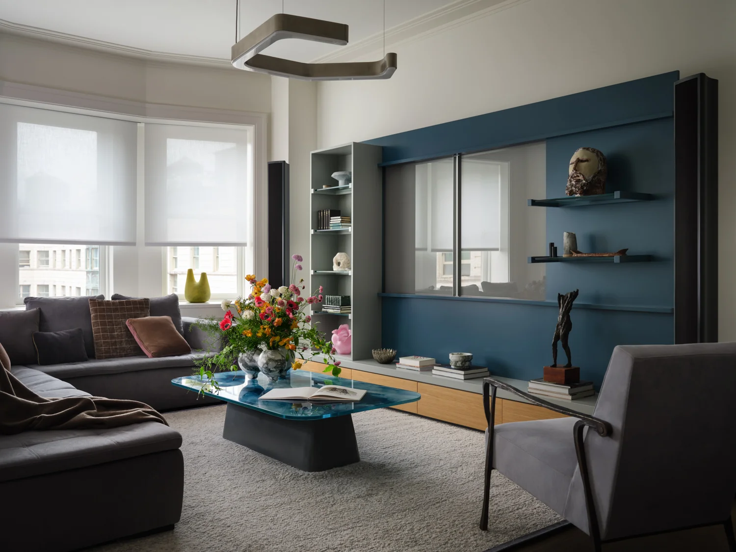 Belnord living room with a gray sectional, blue glass coffee table and blue media unit displaying books and sculptures.