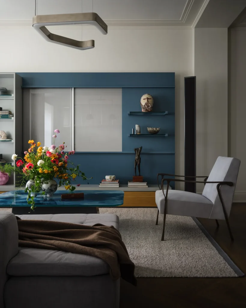 Gray armchair beside a blue coffee table with colorful flowers, backed by shelves displaying collected objects.