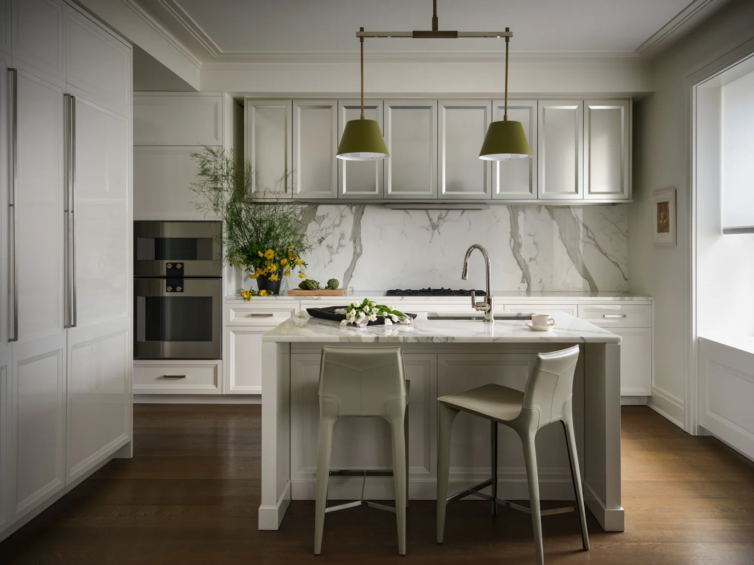 White kitchen with pale counter stools, olive green pendant shades and fresh flowers around a marble island.