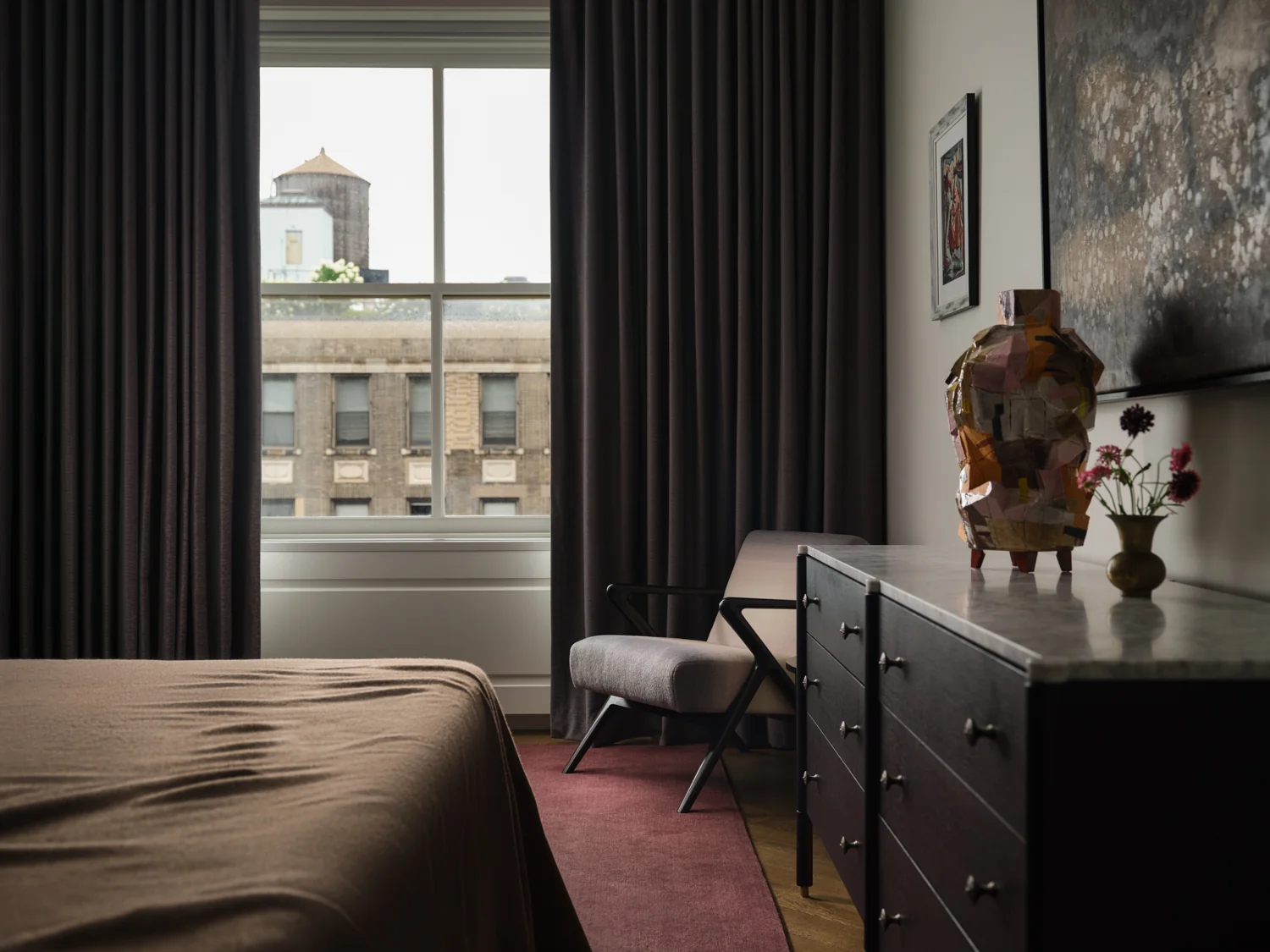 Bedroom corner with a cushioned chair, dark dresser, colorful sculpture and floor-length gray curtains.