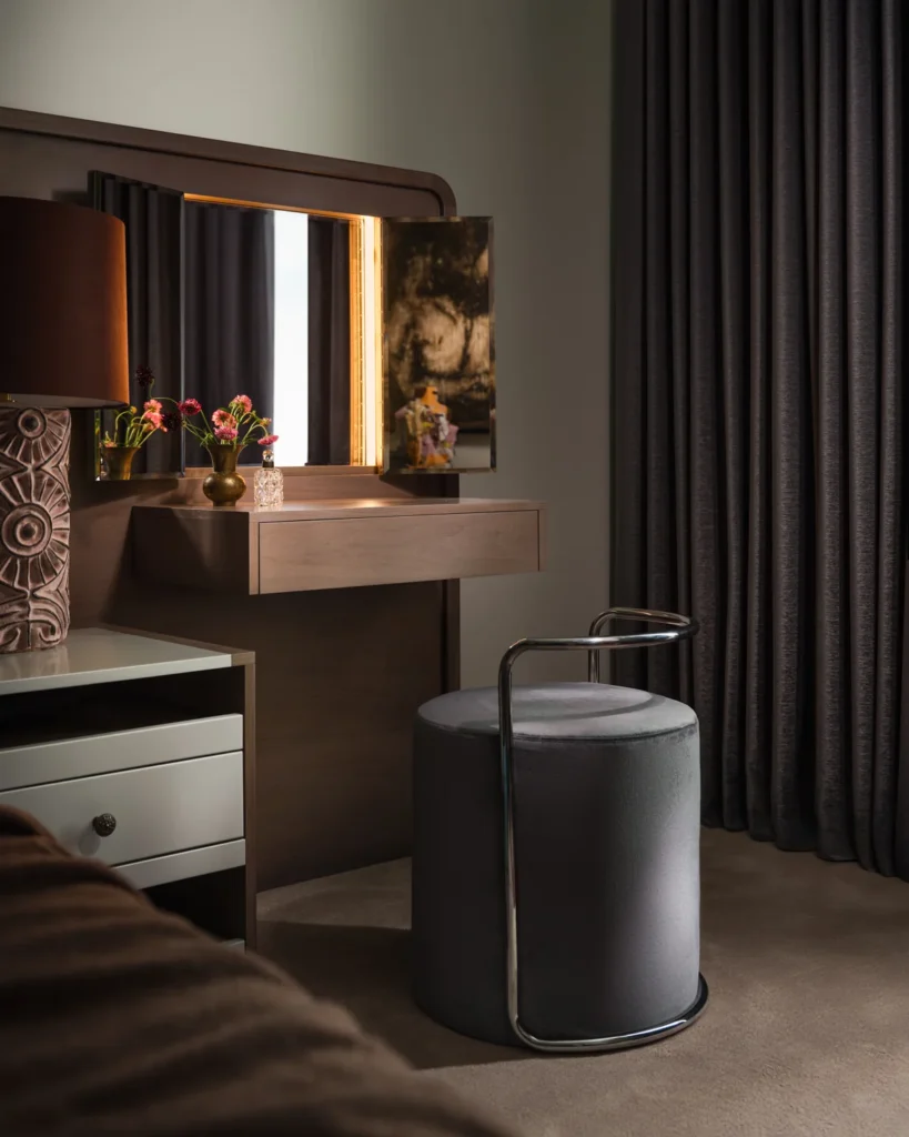 Custom walnut vanity with a lighted folding mirror and round gray stool beside the bed.