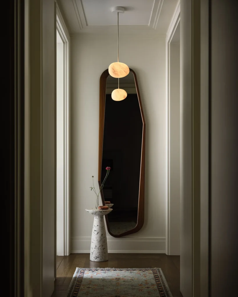 Glowing pendant reflected in a tall hallway mirror above a small stone pedestal table.